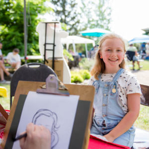 Young girl smiles at the camera as Corrina draws her caricature